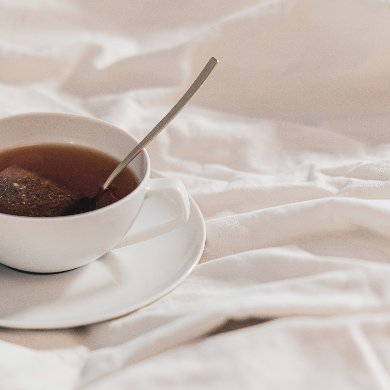 Side close-up of TAC White teacup with saucer, spoon on white sheet.